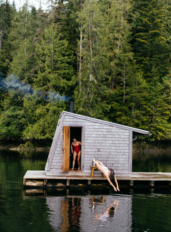 Two people by a small wooden cabin on a dock over water with trees in the background