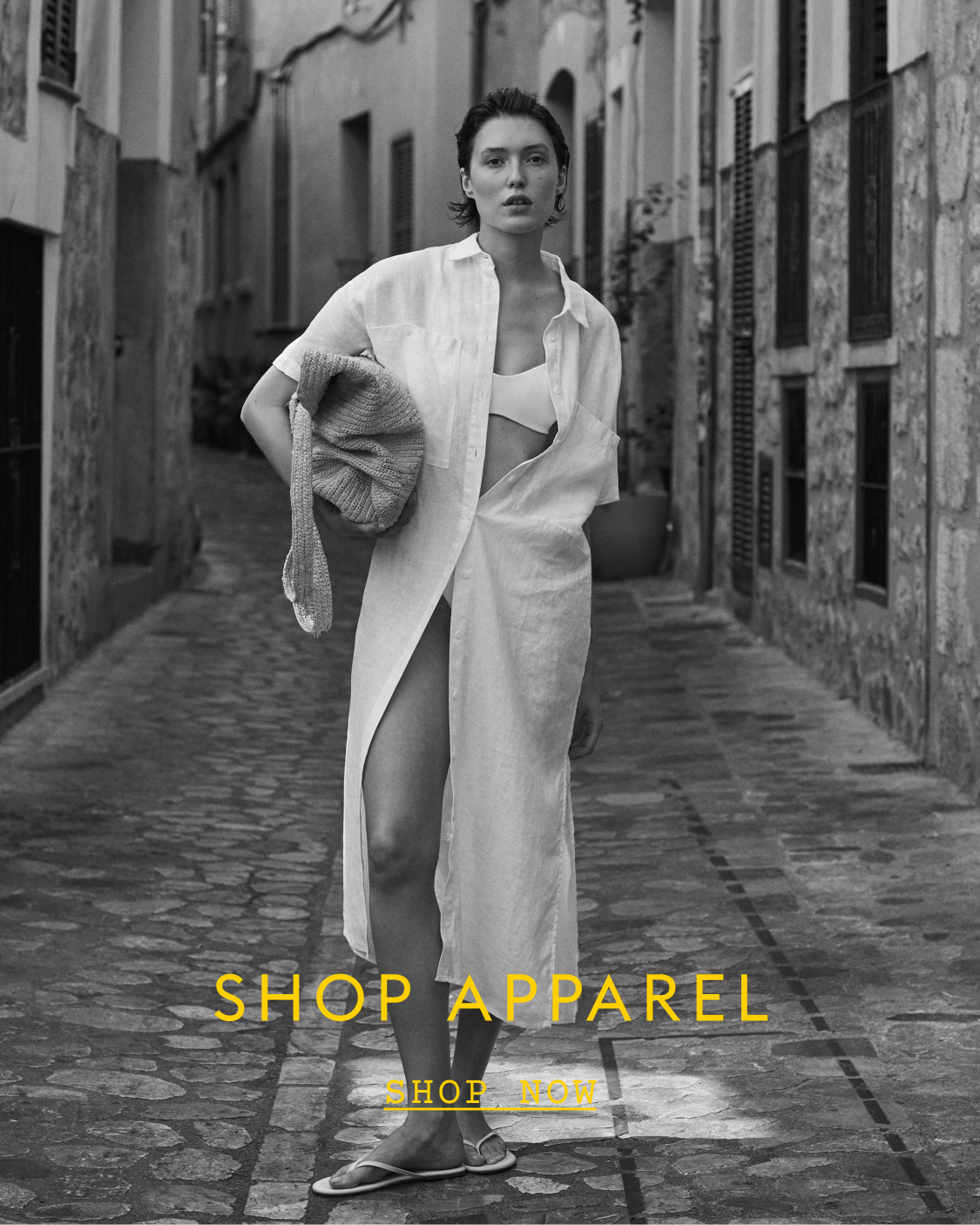 Woman walking down a narrow street wearing a white Getaway Shirt Dress with text 'SHOP APPAREL'.