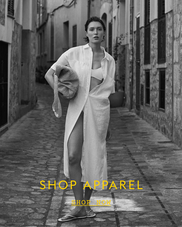 Woman walking down a narrow street wearing a white Getaway Shirt Dress with text 'SHOP APPAREL'.