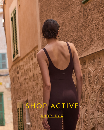 Person wearing a brown Plunge Playsuit in front of a textured wall with 'Shop Active' text.