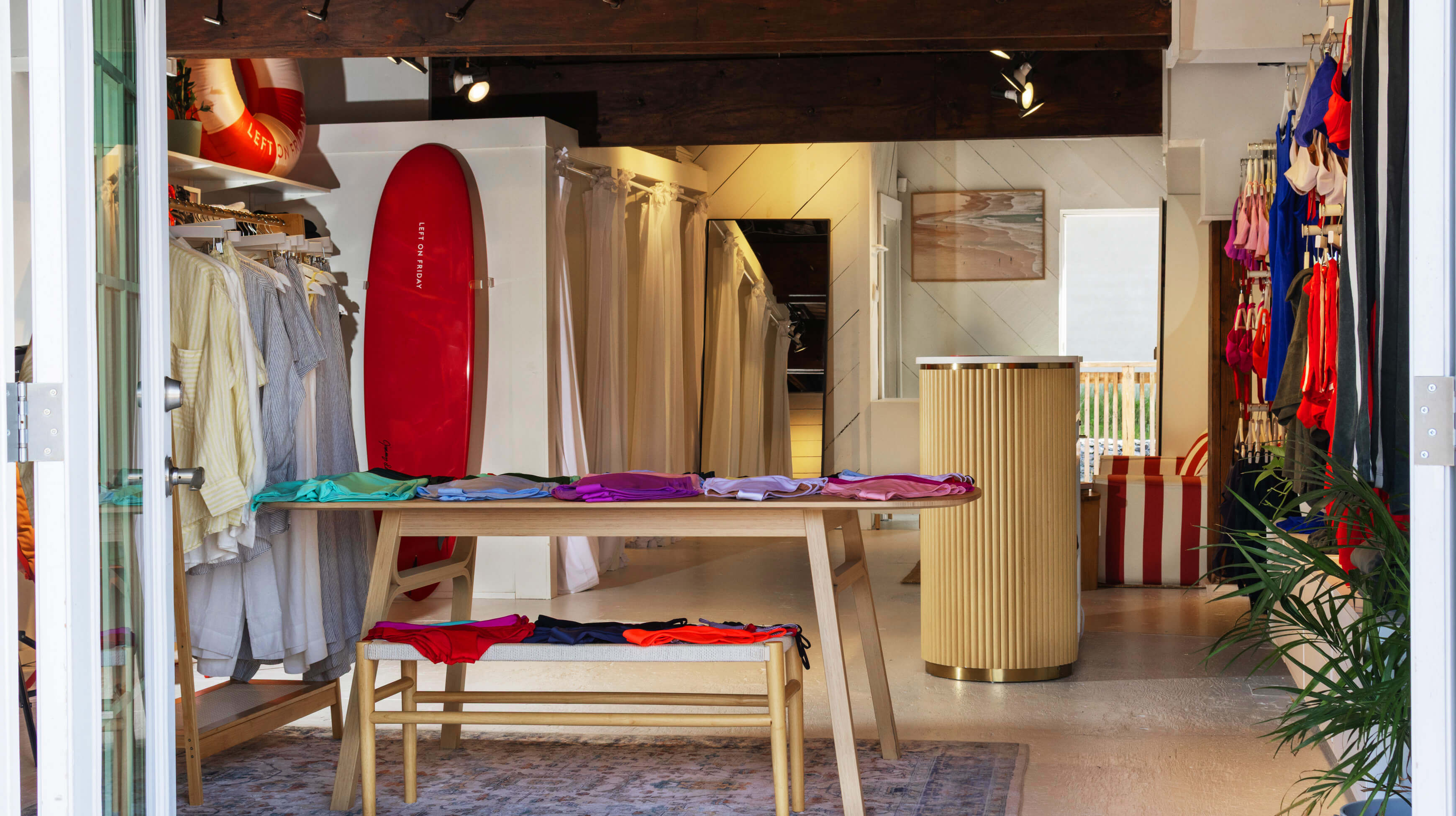 A boutique interior with clothing racks, a wooden table holding folded colorful garments, a bench with folded textiles, a red surfboard leaning against a wall, and a striped bag in the background.