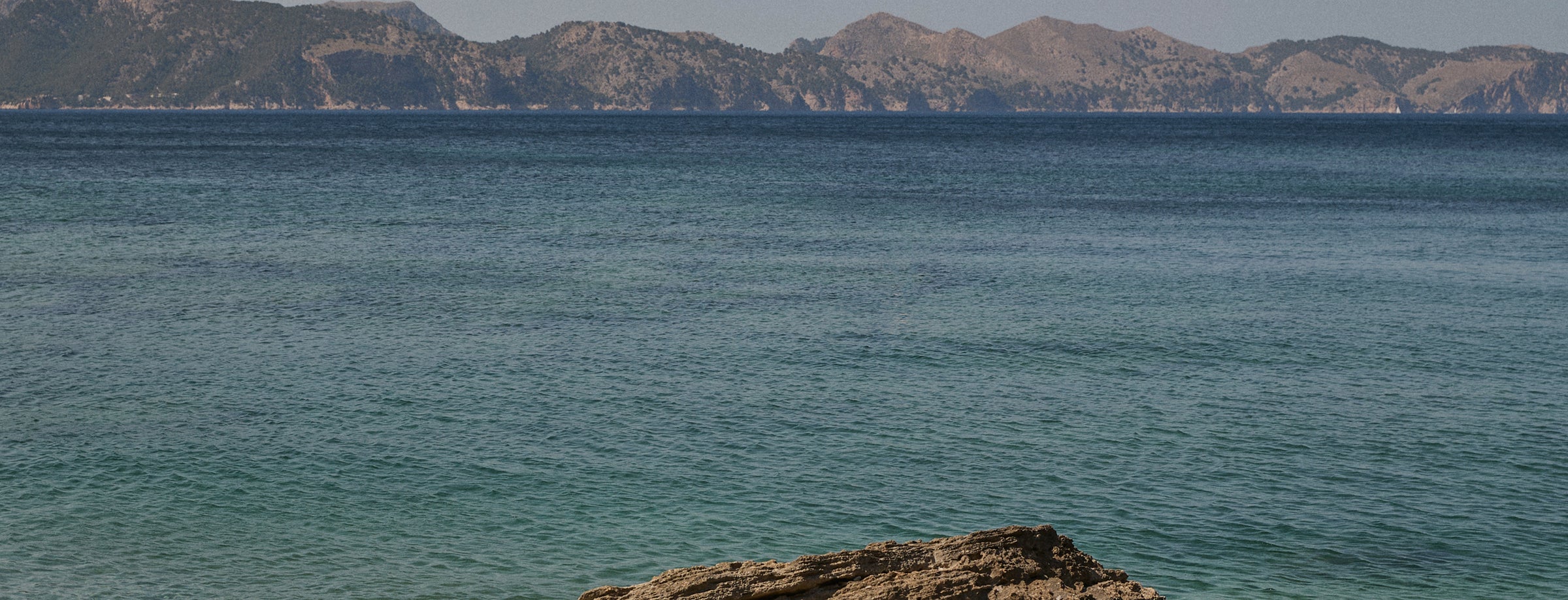 A scenic coastal landscape with calm blue-green water in the foreground, distant rocky mountains, and a pale sky.