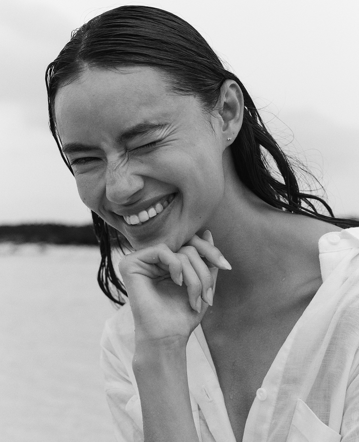 A black and white close-up portrait of a woman with wet hair, smiling broadly and resting her chin on her hand, wearing a white button-up shirt.