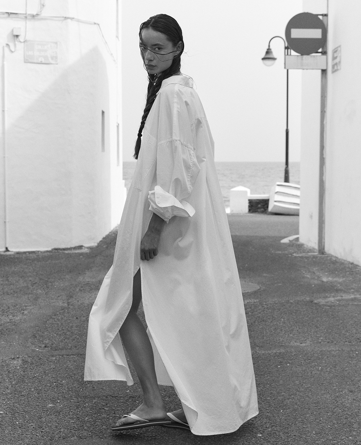 A woman wearing a long white oversized shirt dress walks along a coastal street with white buildings and the sea in the background. The photo is in black and white.