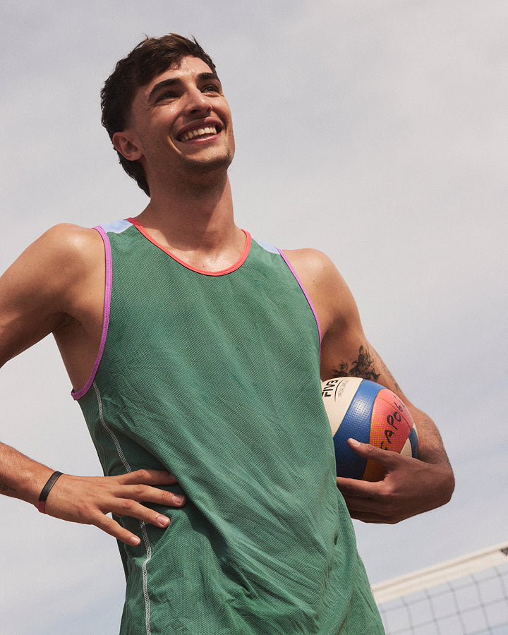 A smiling man wearing a green athletic tank top with pink and purple trim, holding a volleyball outdoors against a cloudy sky.