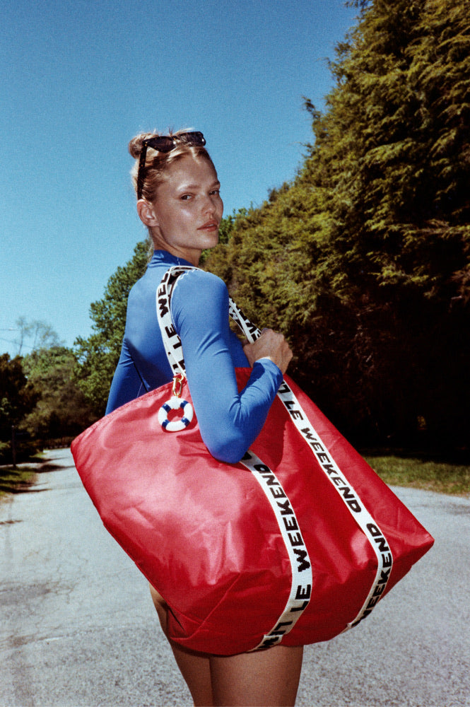 Woman in Rays Long Sleeve Top in First Place holding a large red bag with Le Weekend text on a road with trees in the background