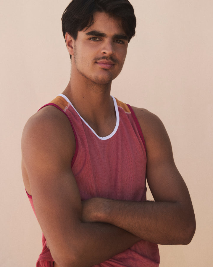A man wearing a sleeveless athletic tank top in dusty rose pink with white and mustard yellow trim, posed with arms crossed against a plain beige background.