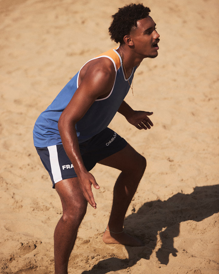 A man wearing a blue and white athletic tank top and navy shorts with white side stripes is crouching on a sandy beach. The brand name 'Celsino' is visible on the thigh of his shorts.
