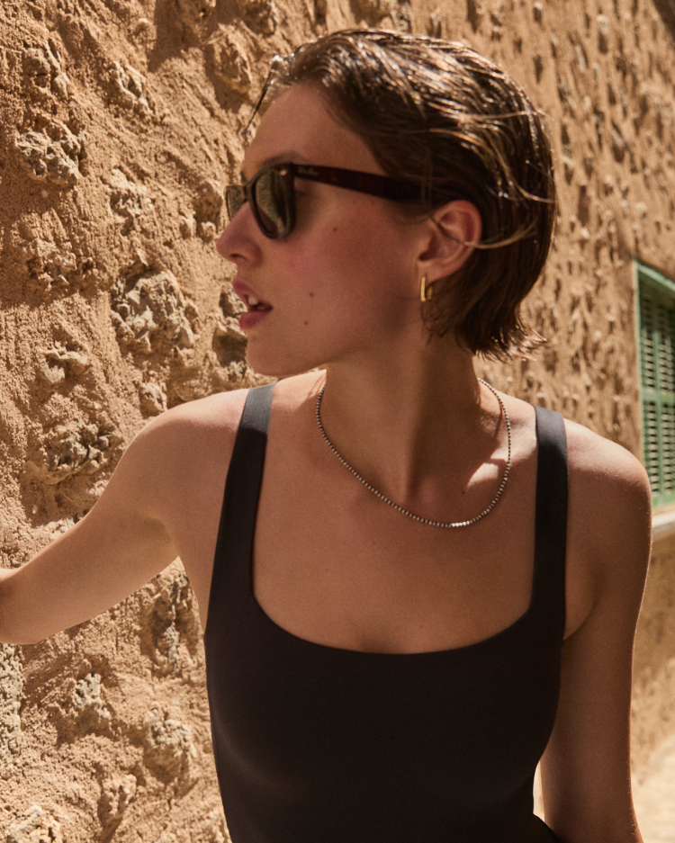 Woman in a black Streamline Suit and sunglasses standing against a textured stone wall.