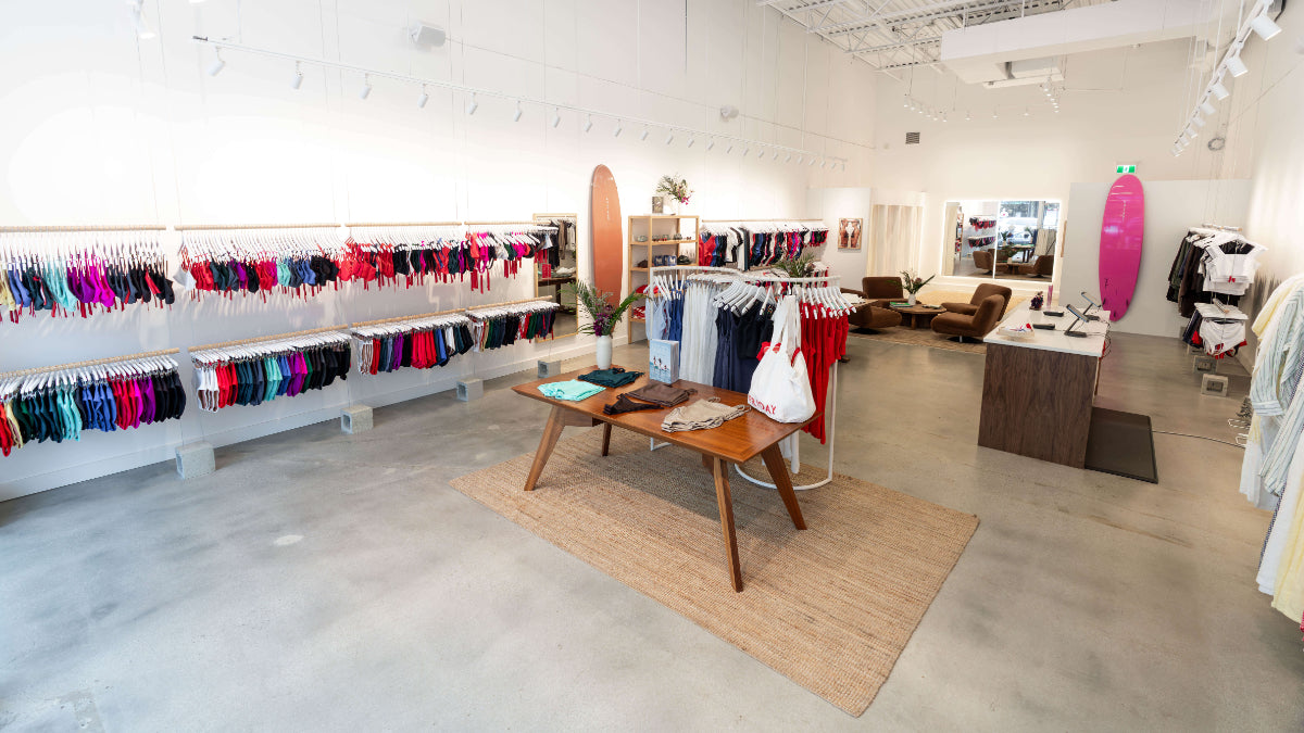 A wide, bright boutique interior showing racks of colorful swimwear and apparel, a central wooden table with folded garments, a beige rug, concrete floors, and surfboard decor on the wall.