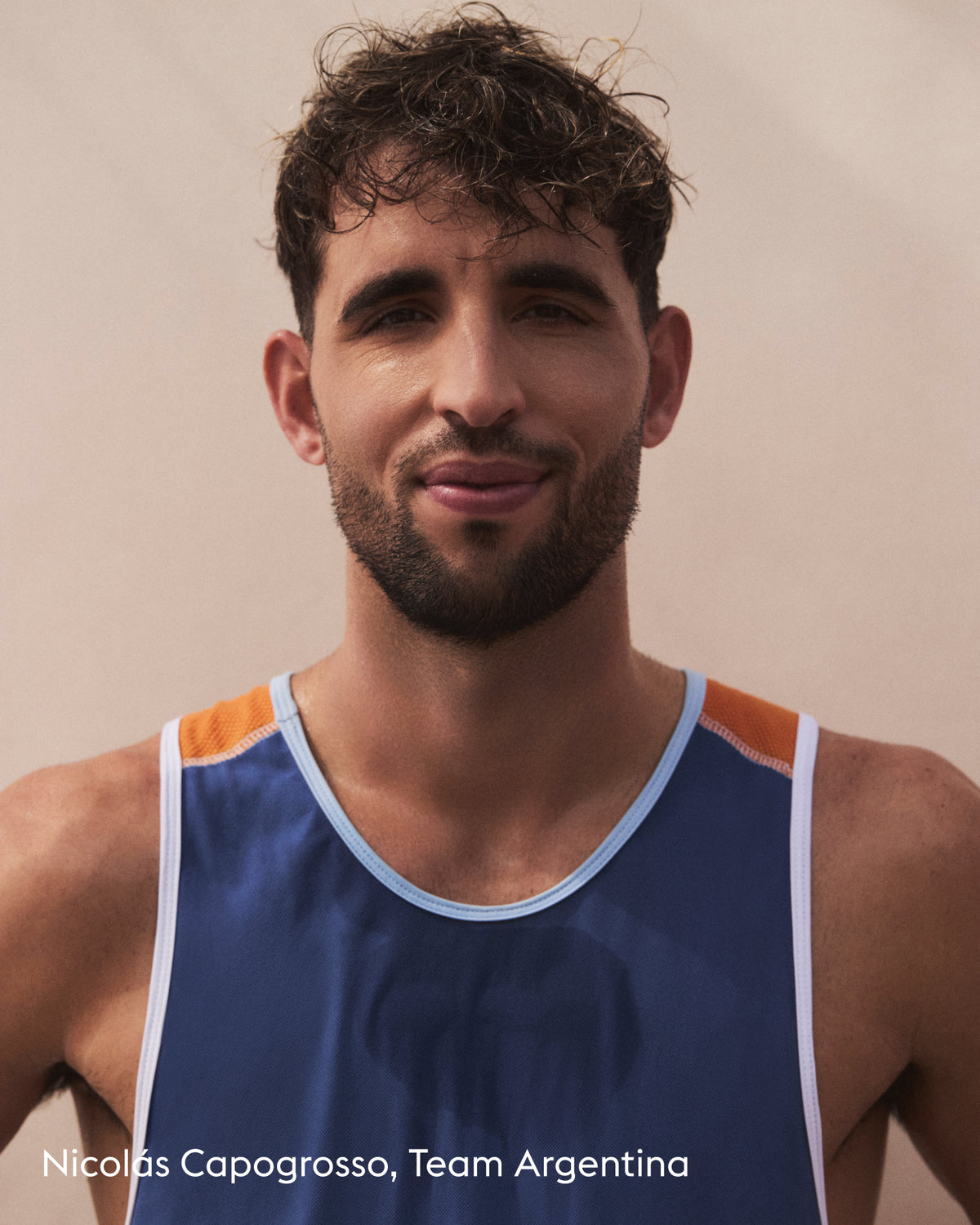 A close-up portrait of a man wearing a blue athletic tank top with orange shoulder panels and white trim, against a plain beige background. White text at the bottom reads 'Nicolás Capogrosso, Team Argentina'.