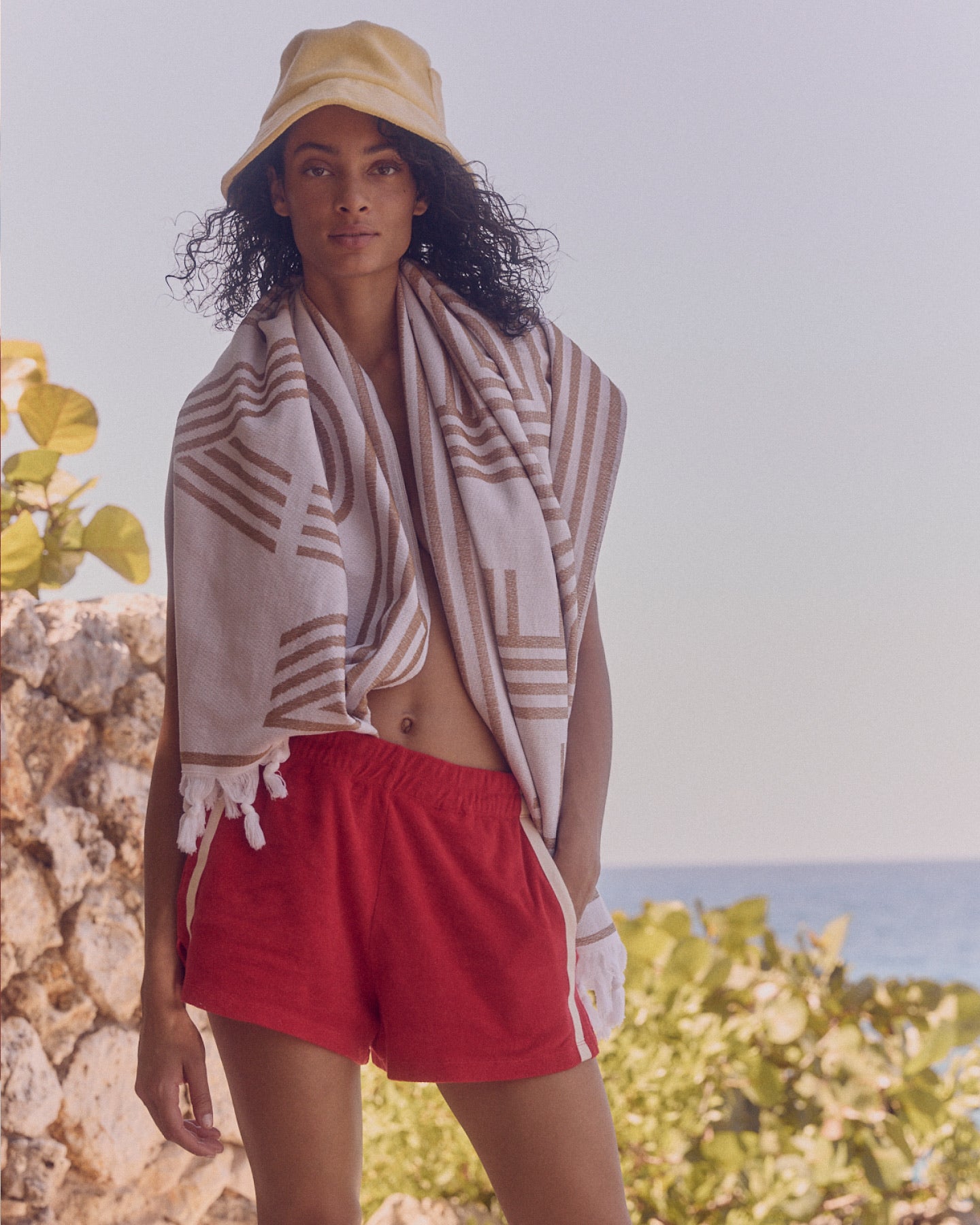 A woman outdoors wearing a beige bucket hat, a cream and brown striped fringed towel draped over her shoulders, and bright red shorts, with a stone wall and blue sea in the background.