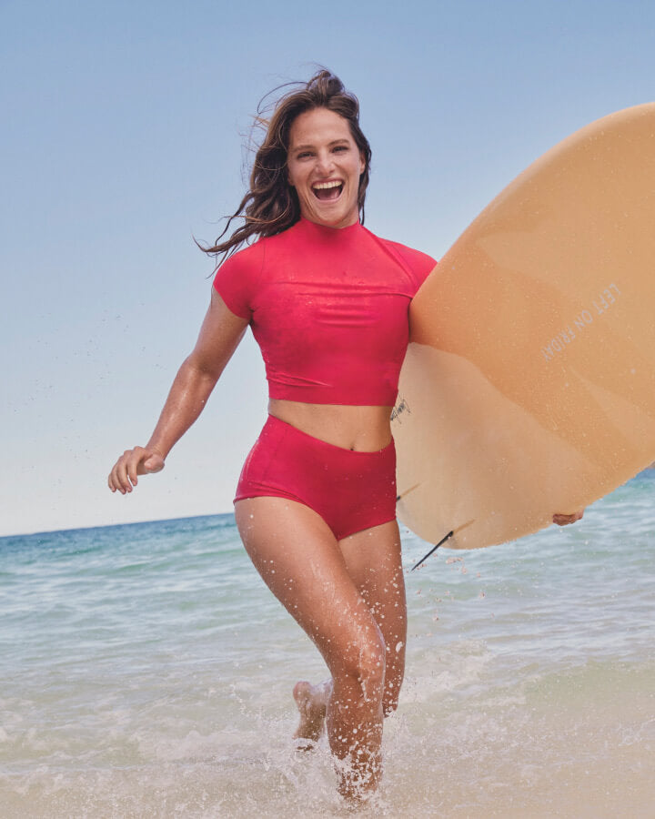 A woman running through shallow ocean water carrying a large beige surfboard, wearing a Rays Short Sleeve Top in Rescue with the Barrel Bottom in Sweet Chili Heat.