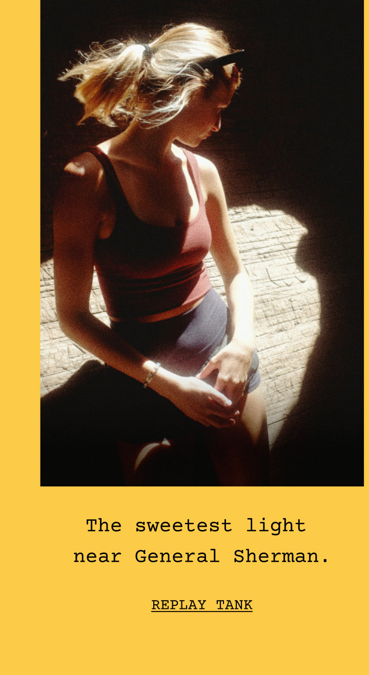 A woman wearing the replay tank in jam and ace short in jet shown from behind in a forest setting, with a yellow border and text below reading 'En route to visit General Sherman.' and 'REPLAY TANK.