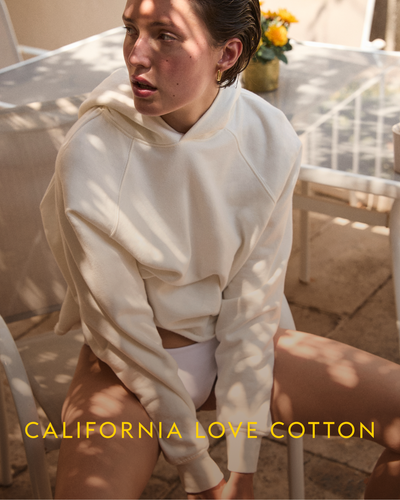 Person wearing a cream Homerun Hoodie sitting outdoors with 'California Love Cotton' text.