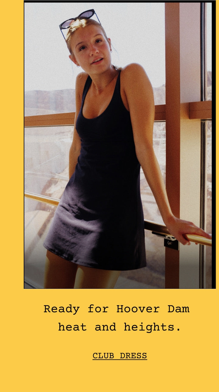 A woman wearing The Club Dress in Jet stands by a window, with text below reading 'Ready for Hoover Dam heat and heights.' and 'CLUB DRESS' on a yellow background.