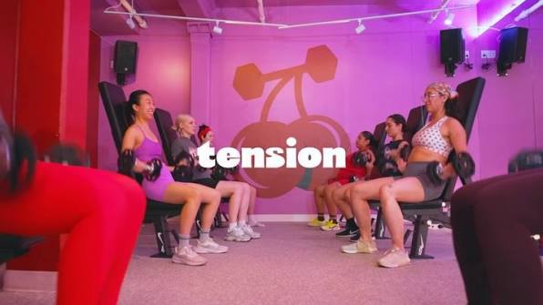 A group of women exercising on seated cable machines in a fitness studio with pink walls and a large dumbbell-and-cherries graphic on the back wall. The word 'tension' is overlaid across the center.